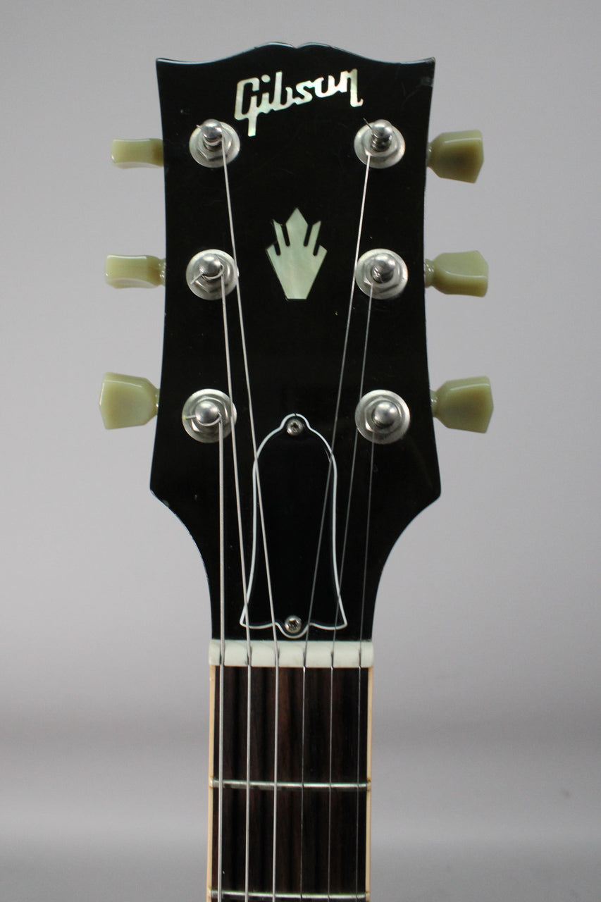 Close-up of the black headstock with a mother-of-pearl crown inlay, six cream tuning pegs, and a bell-shaped truss rod cover.