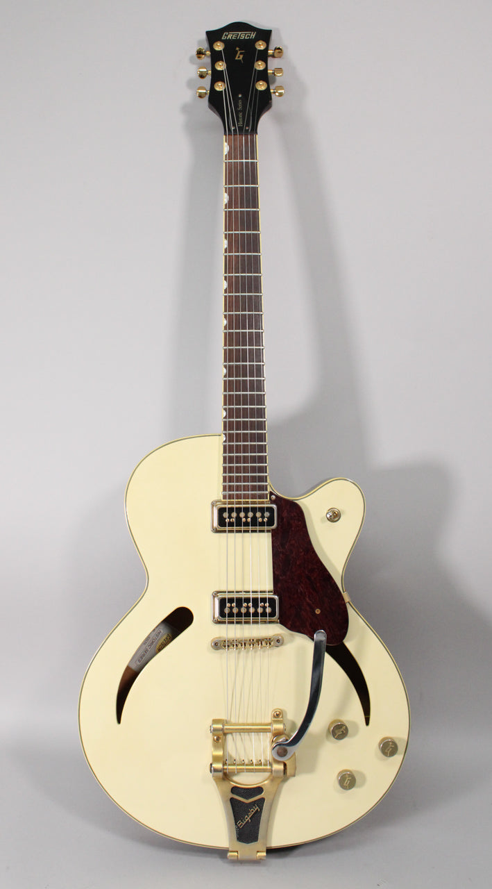 Full-length, front view of an antique white hollow-body electric guitar with gold hardware, f-holes, and a Bigsby vibrato.
