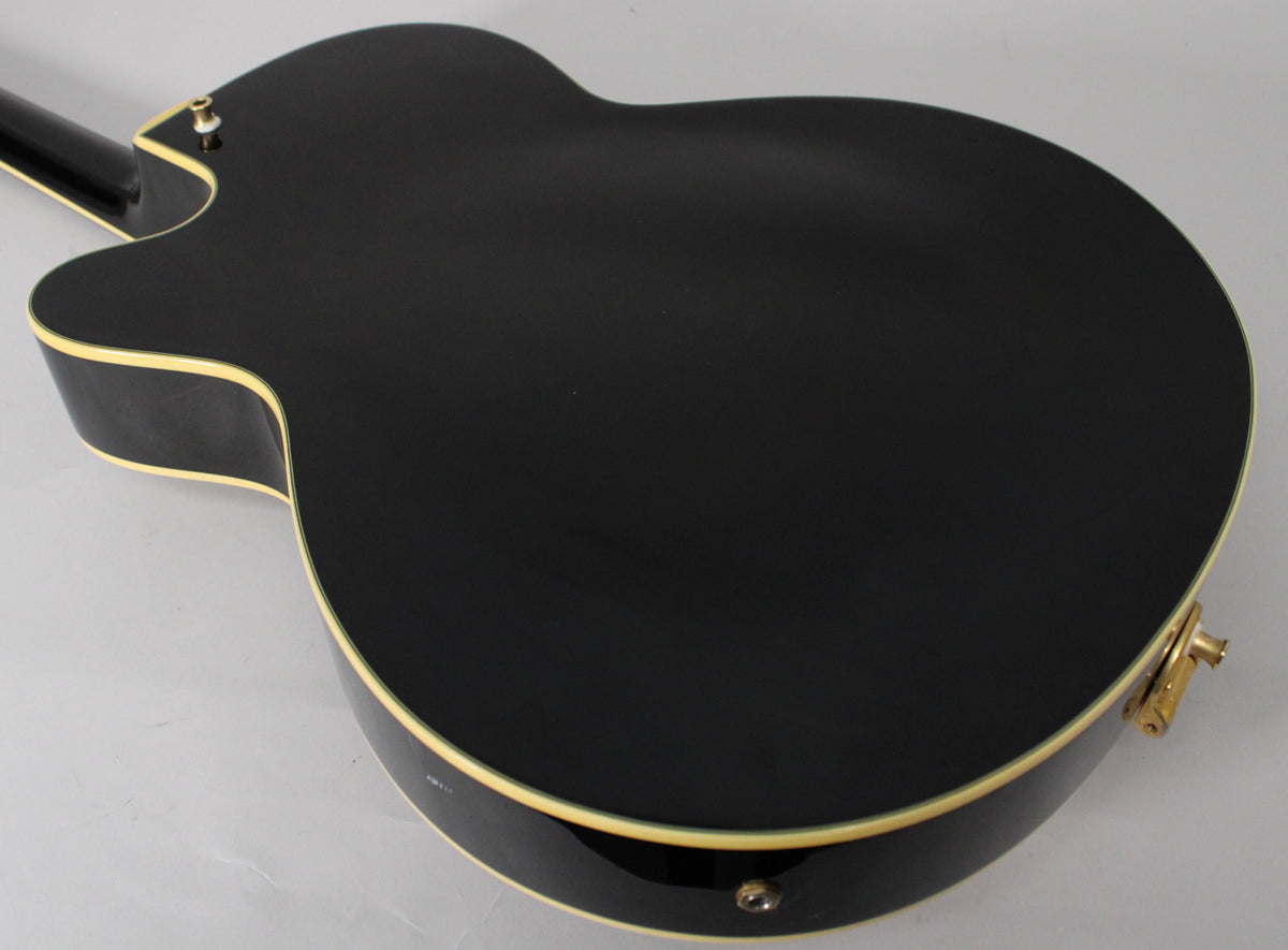 The guitar's glossy black back and sides are visible, with cream binding, a gold strap button, and an input jack.