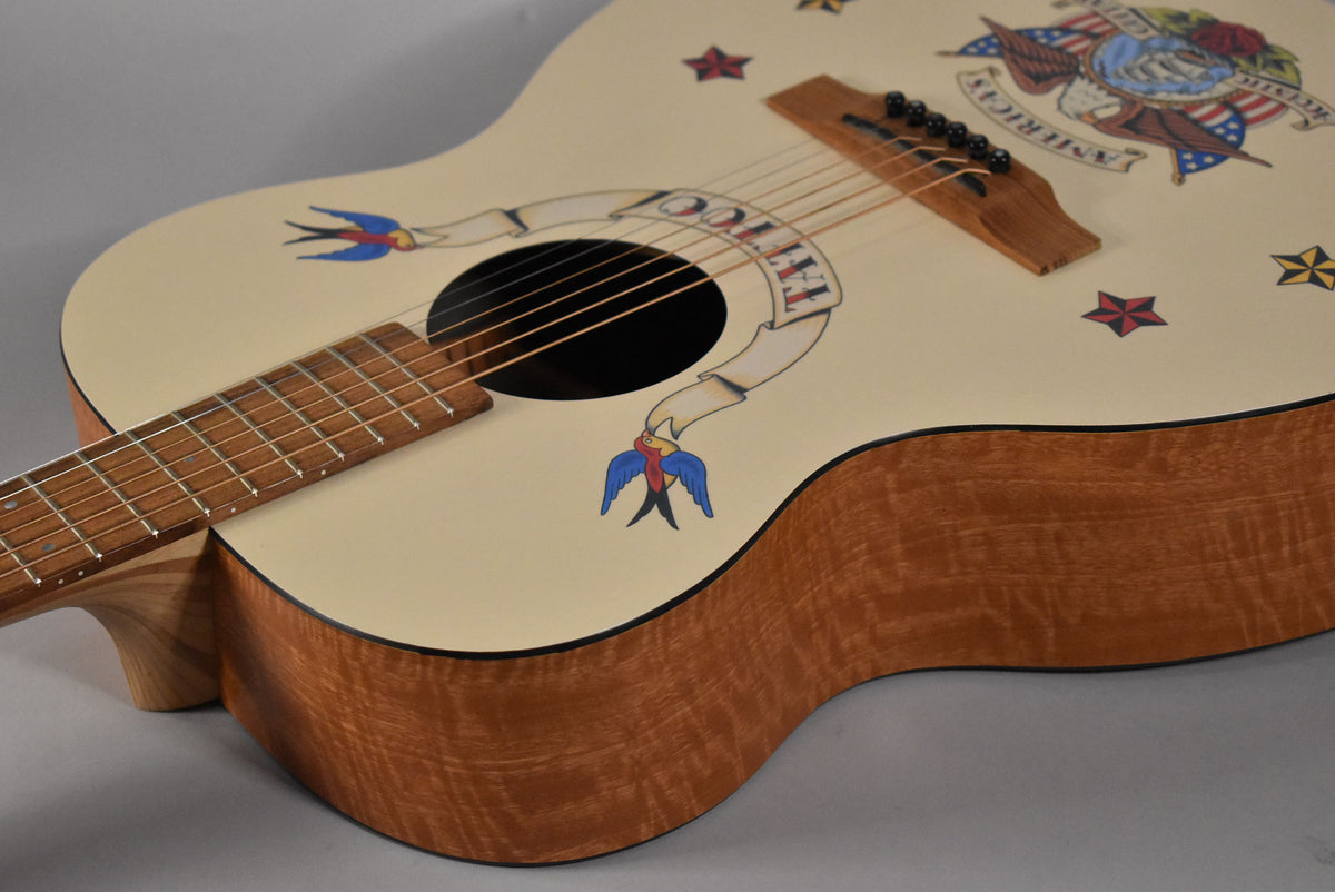 The cream-colored acoustic guitar body, with a tattoo-style banner and swallows, shows its figured wood sides and fretboard from above.