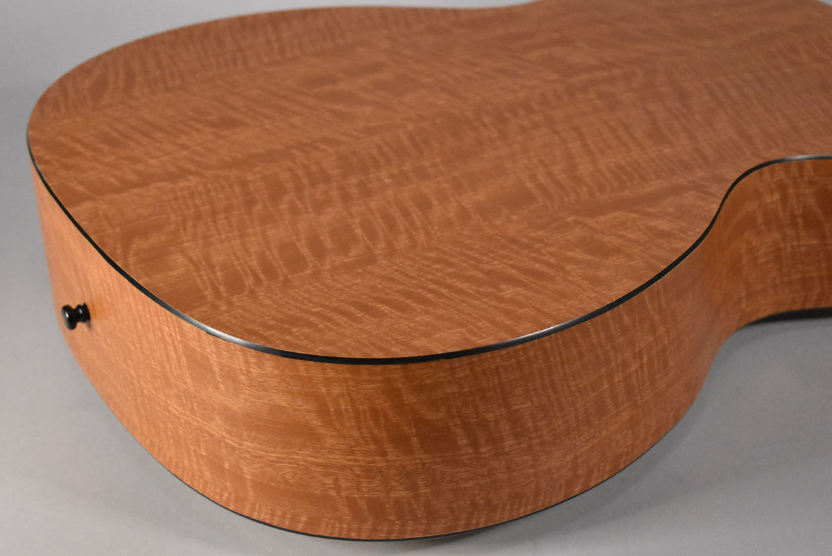 The lower bout and side of the acoustic guitar, showing the light brown wood grain with a wavy, figured pattern and black binding.