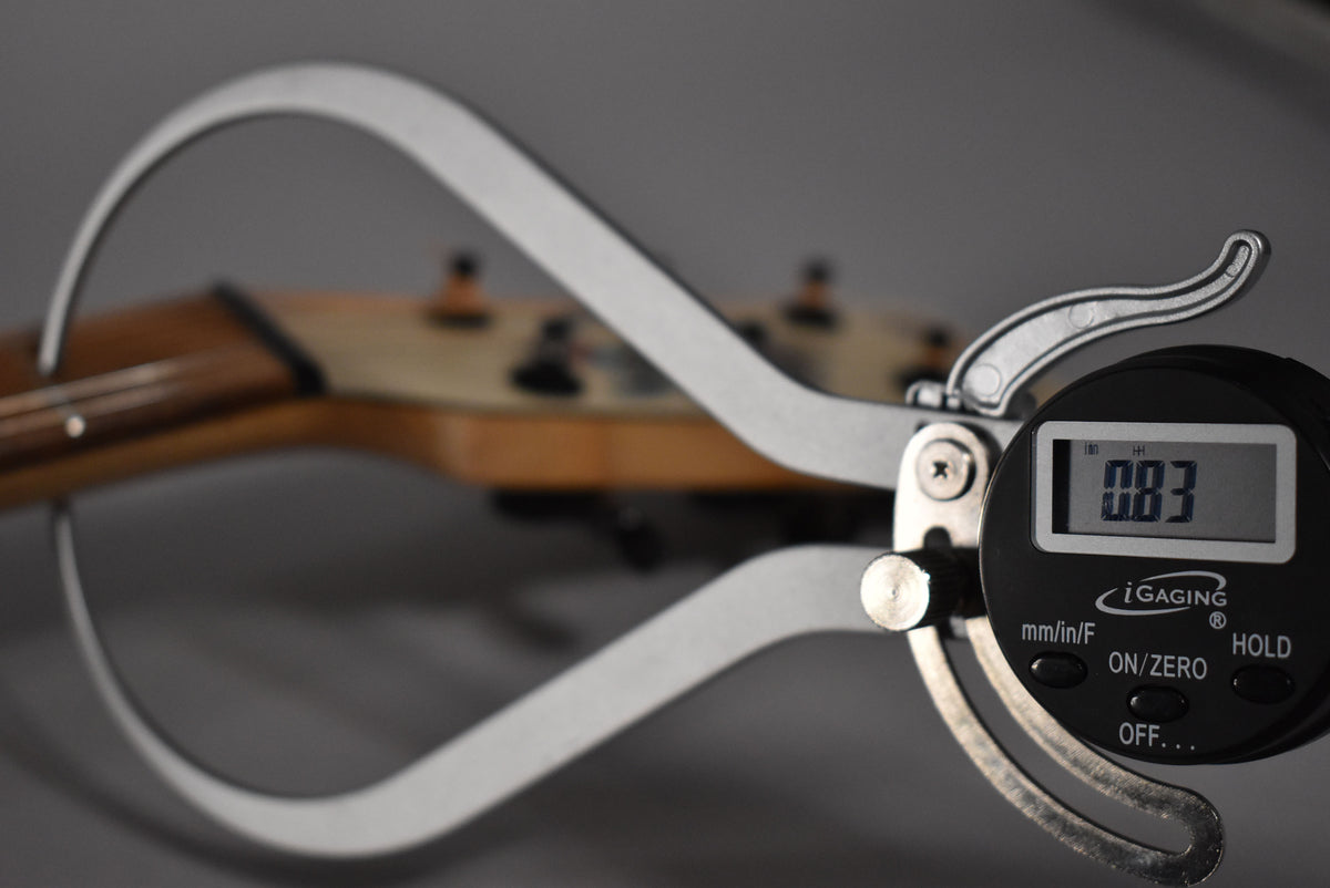 A digital caliper measuring the guitar's headstock, displaying "0.83" on its screen, with tuning pegs blurred in the background.