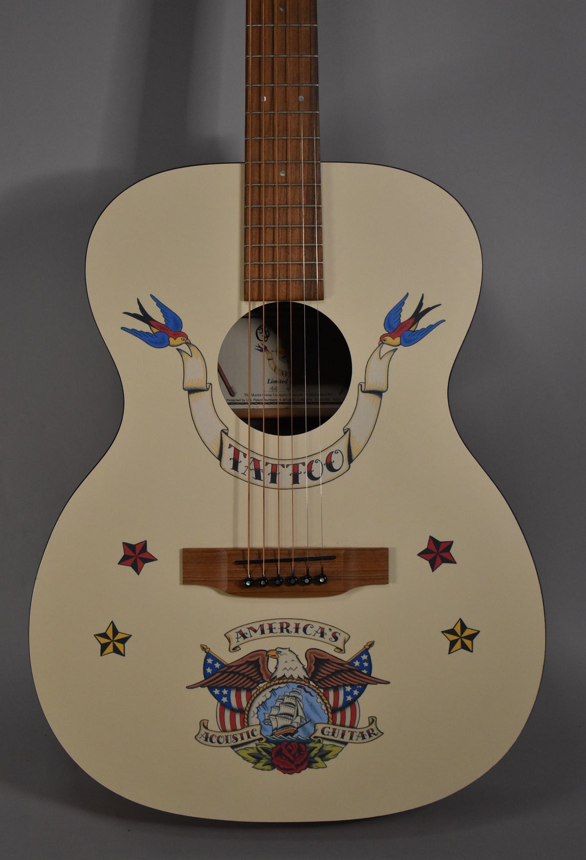 The acoustic guitar's cream body is adorned with colorful traditional tattoo art, including swallows, stars, and an eagle.