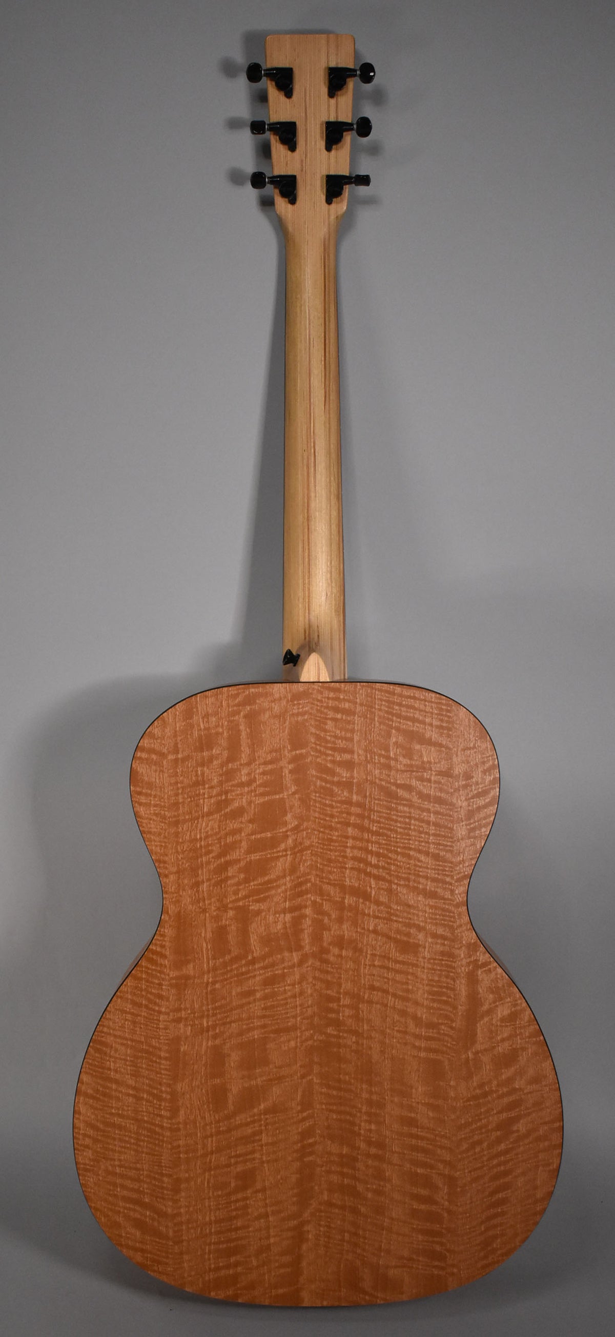 Full rear view of the acoustic guitar, showing a light brown figured wood body, a lighter plain wood neck, and black tuners.