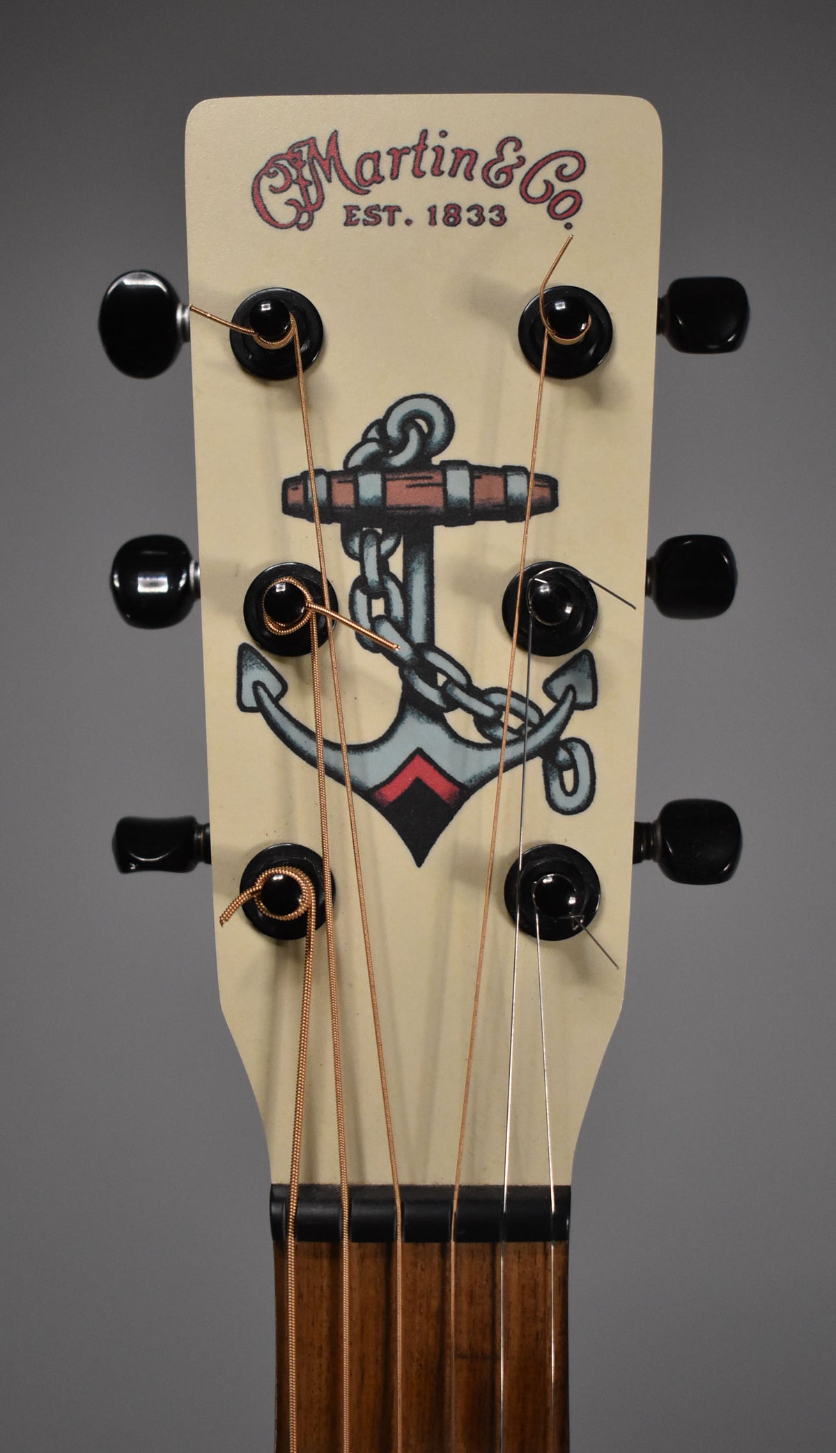 Close-up of a guitar headstock with "Martin & Co. EST. 1833" and an anchor tattoo graphic, six tuning pegs, and strings.