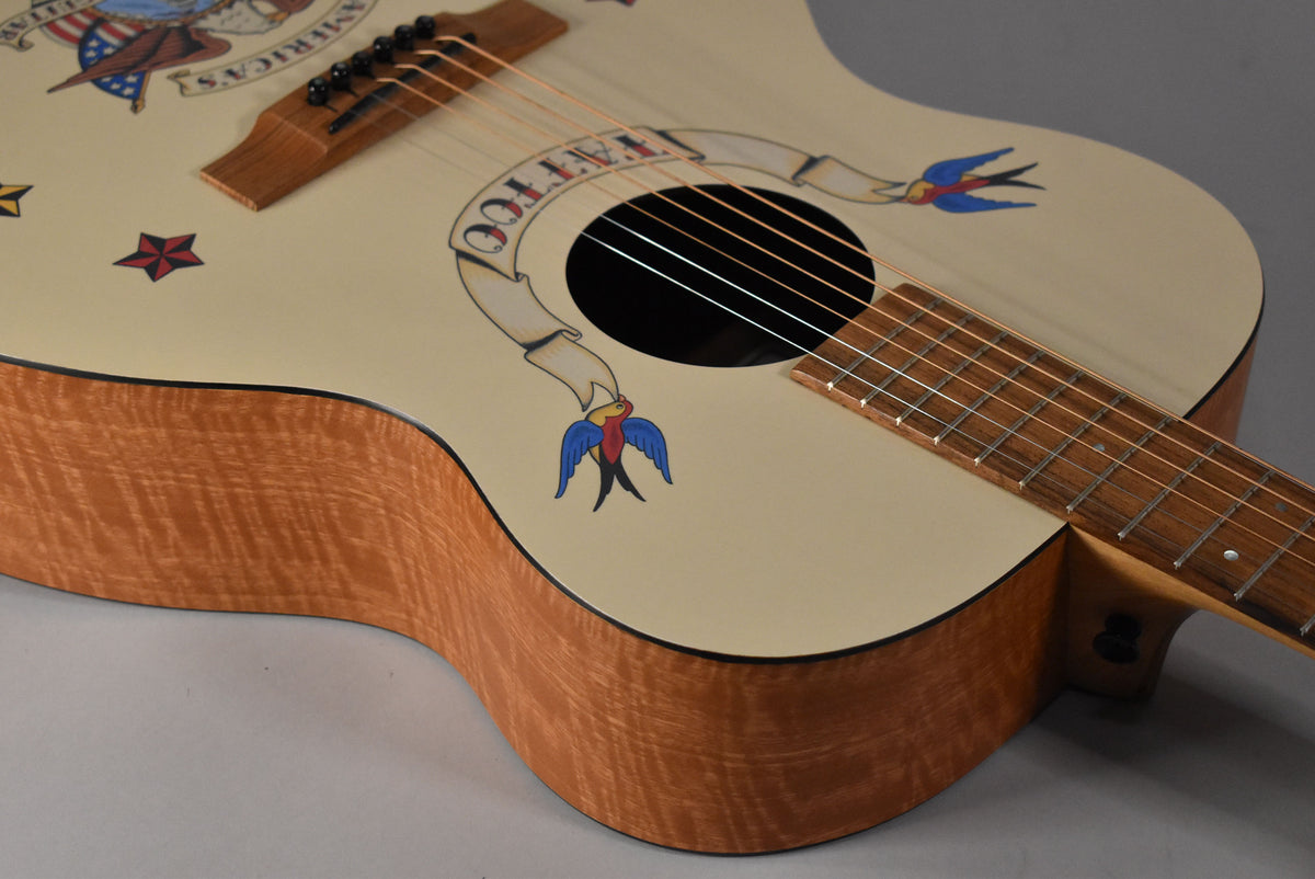 The acoustic guitar's cream top features colorful tattoo-style art, with a banner around the soundhole and a swallow below it.