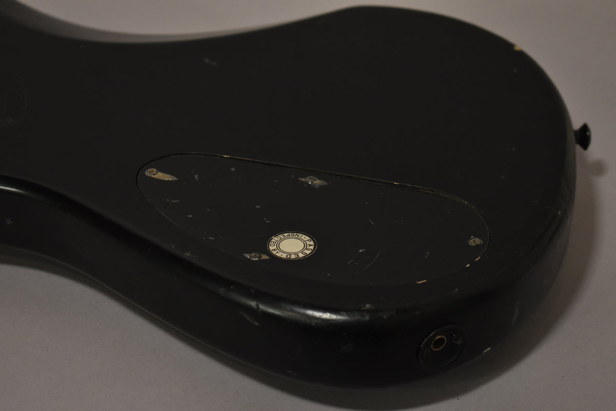 Close-up of the matte black bass guitar's lower back, showing a control cavity cover with a round white sticker and an output jack.