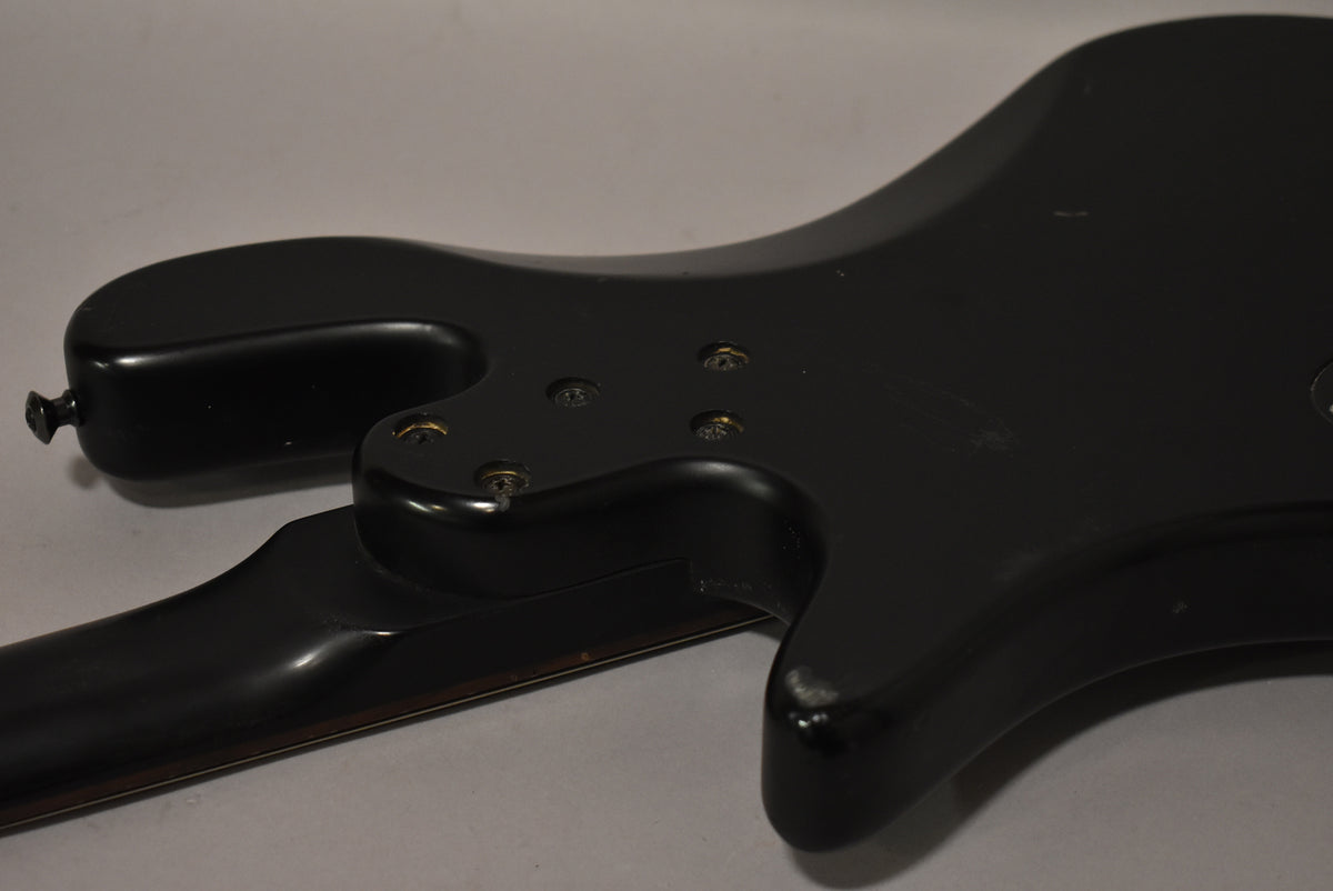 The matte black bass guitar's body and neck joint are viewed from the back, showing four neck screws and minor scuffs.