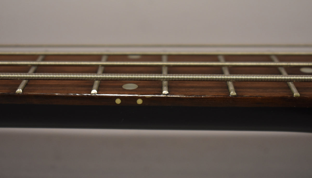 Close-up of the bass fretboard, showing four strings, frets, dot inlays, and wear along the dark brown edge.