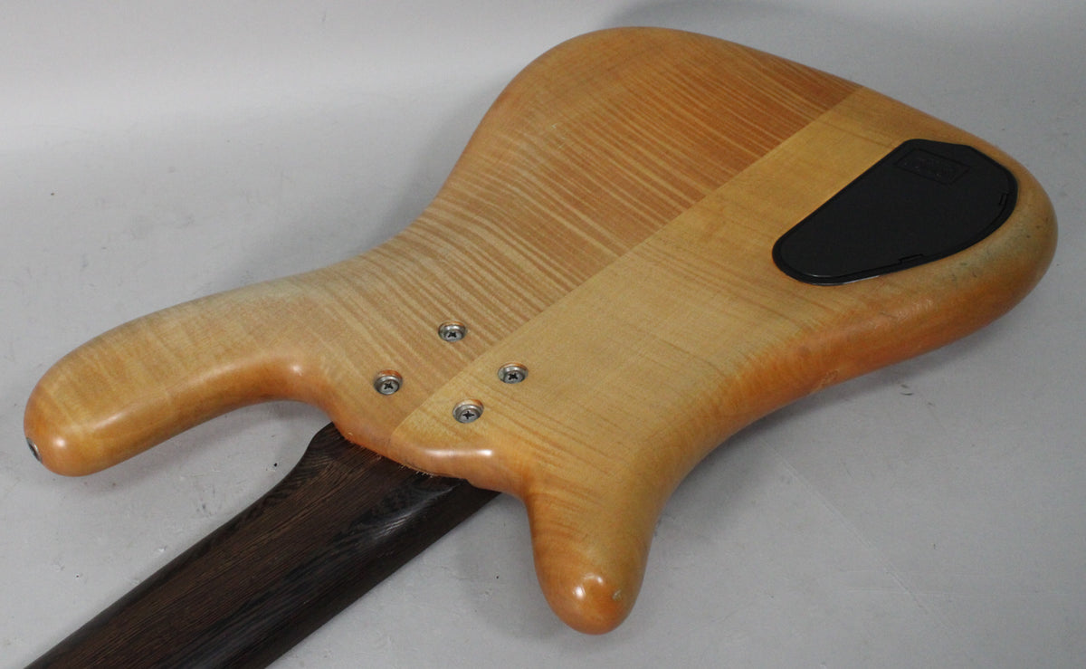 The bass's natural transparent satin finish body, with a visible wood grain, shows a black battery compartment and four neck screws.