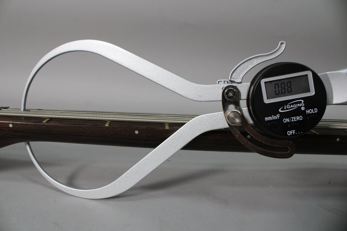 A digital caliper measures the bass's dark wood fretboard and strings, displaying "0.88" on its screen.