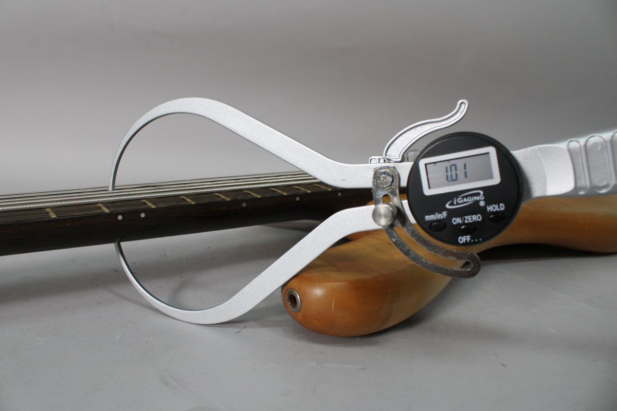 A digital caliper measures the bass's dark fretboard and strings, showing "1.01" on its display.