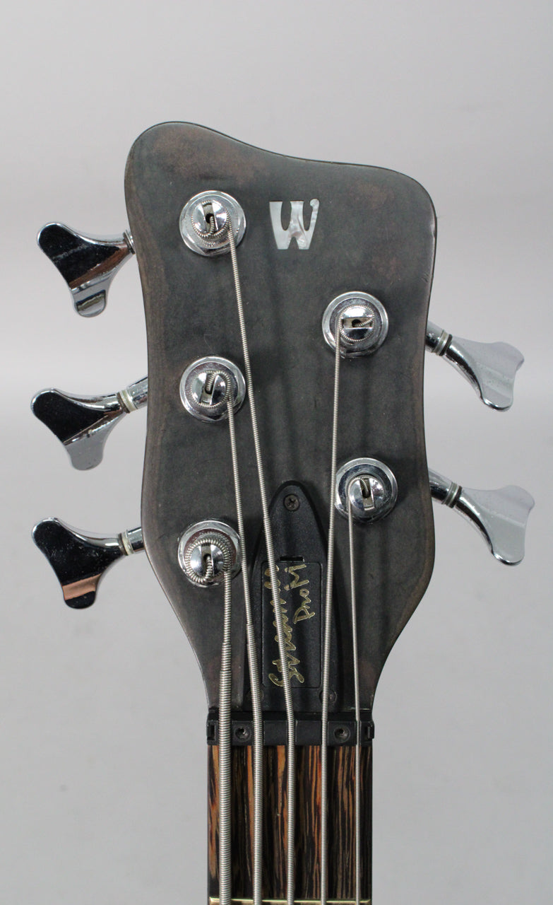 The bass headstock, dark natural wood with a satin finish, shows five chrome tuners and a striped fretboard.