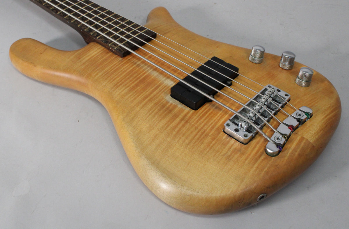 The bass body, angled from the neck, shows a natural transparent satin finish with flame maple grain, black pickups, and chrome hardware.