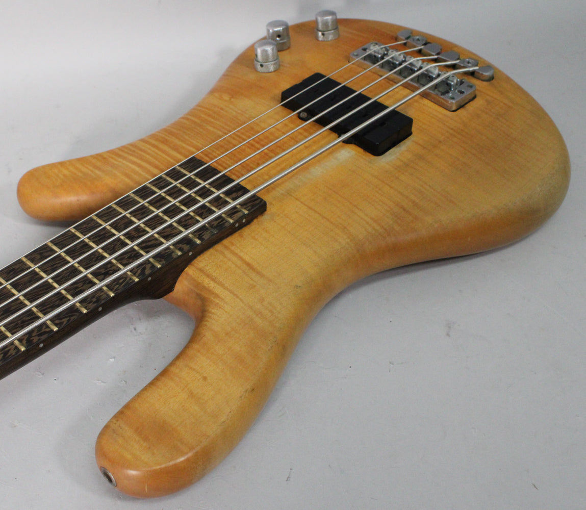 The bass body, angled from the neck, shows a natural transparent satin finish with flame maple grain, black pickups, and four silver control knobs.