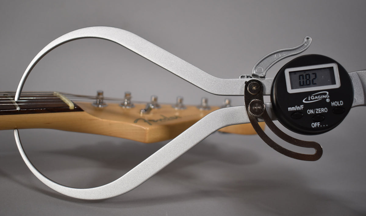 A digital caliper measures the guitar's neck relief at 0.82, with the headstock and tuning pegs visible in the background.