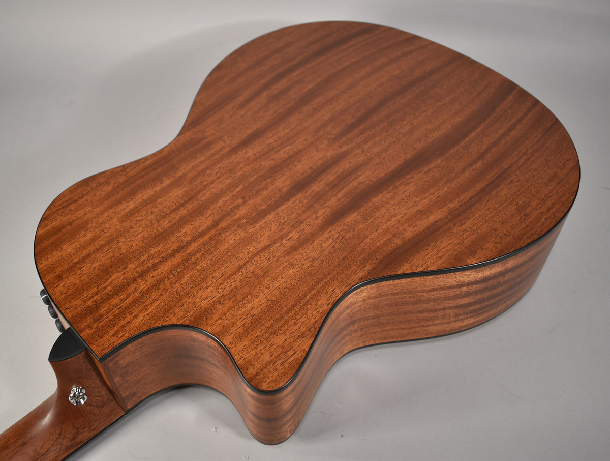 The back of a natural finish acoustic guitar, showing the mahogany wood grain and black binding, with a strap button visible.