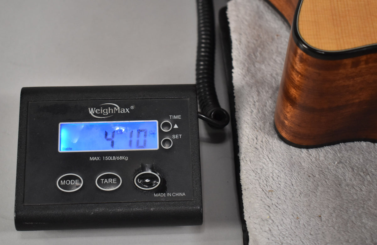 A digital scale displays "4 lb 10 oz" next to the lower bout of the natural finish acoustic guitar, resting on a white towel.