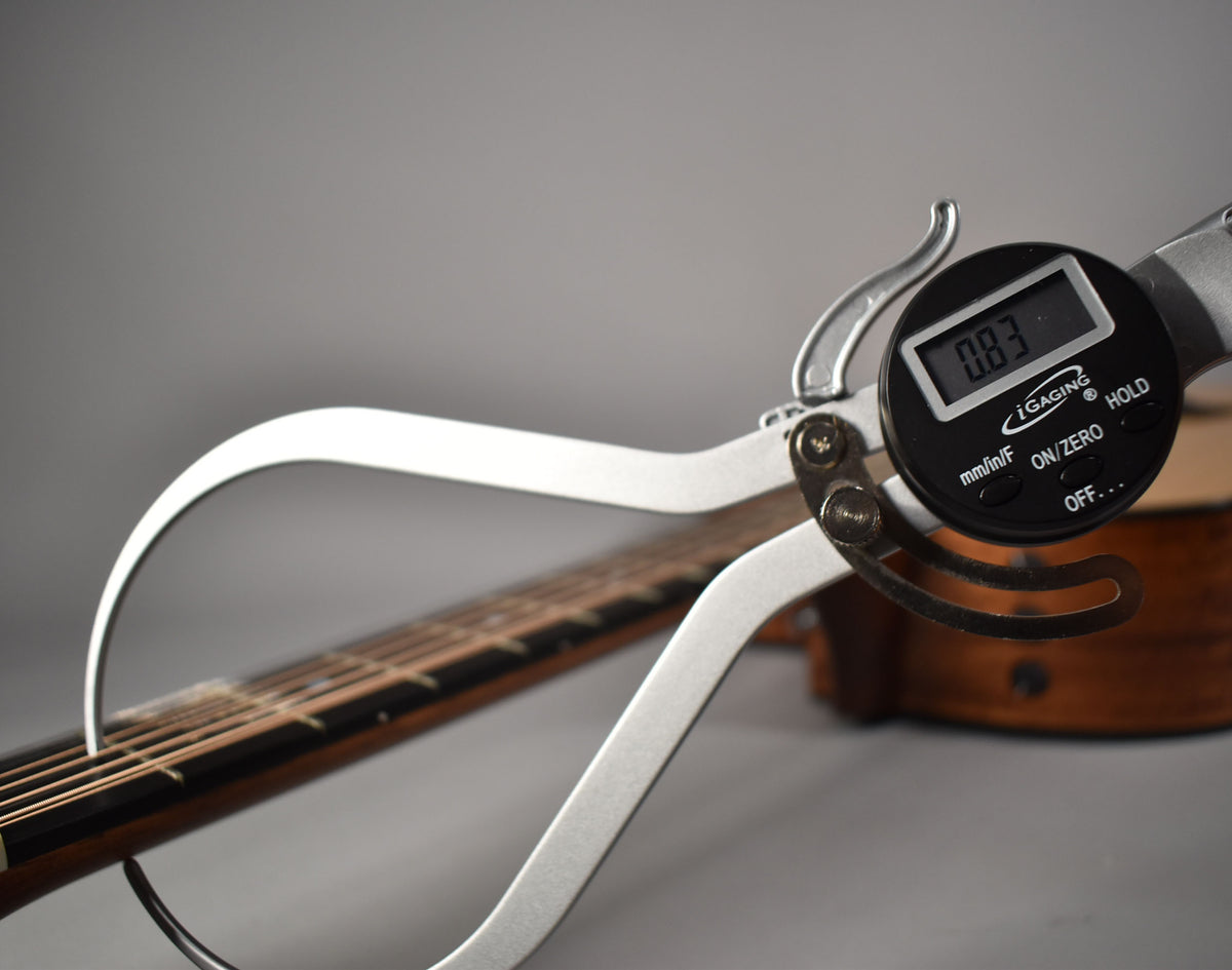 A digital caliper measures the guitar's natural finish neck, showing a reading of 0.83, with the body blurred in the background.