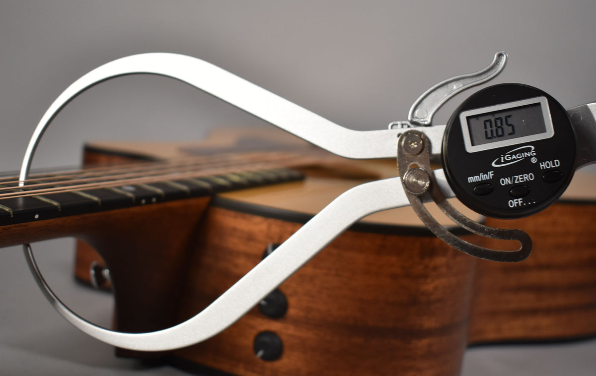 A digital caliper measures the guitar's action at the 12th fret, showing 0.85, with the natural wood body visible.