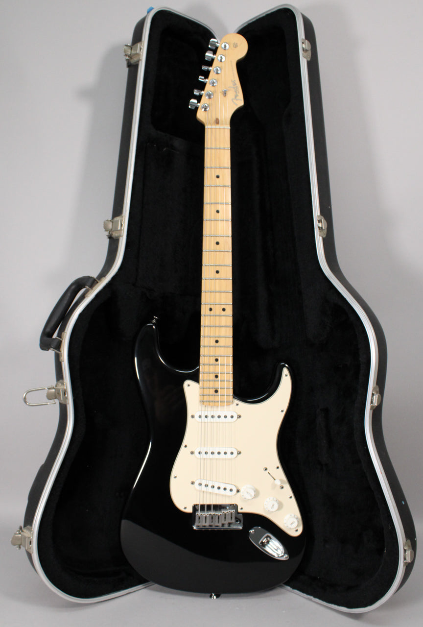 A black electric guitar with a maple neck and white pickguard, resting in a black hard shell case, viewed from the front.