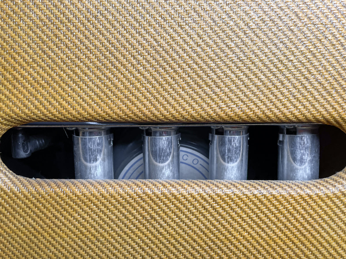 Close-up of the tweed amp's rear, showing four clear vacuum tubes and a glimpse of a speaker cone through the open back.