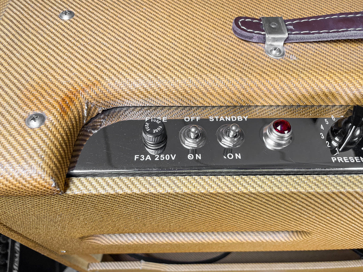 Close-up of the tweed amp's top right, showing the brown leather handle, a dark control panel with fuse, power switches, and a red jewel light.