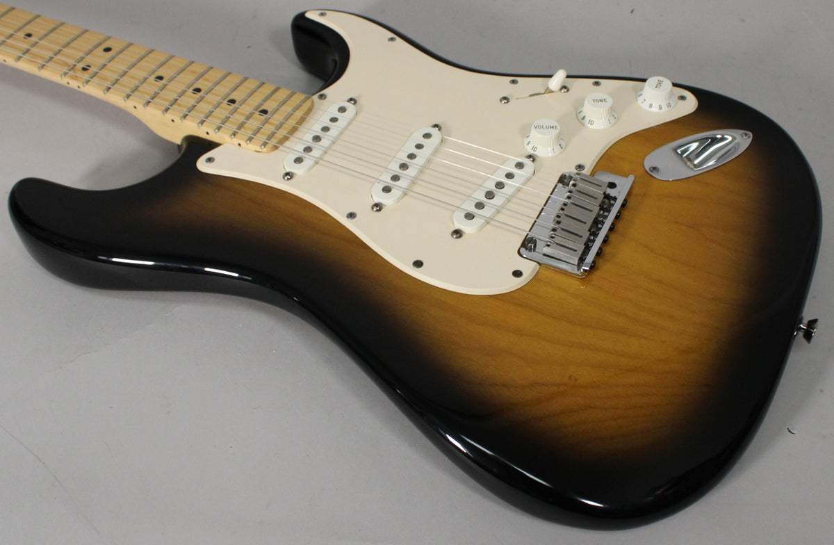 Angled view of a sunburst Stratocaster, showing the body, cream pickguard, three single-coil pickups, and maple neck with dot inlays.