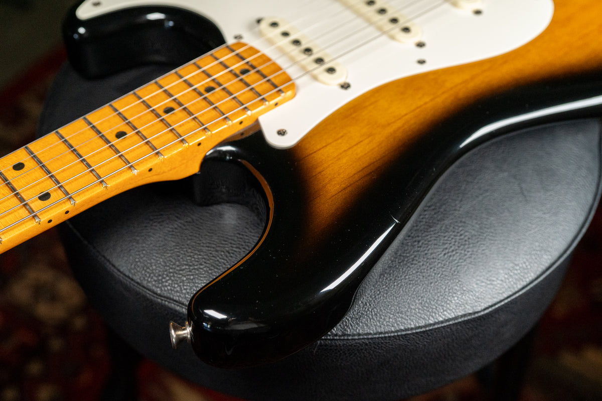 The guitar's lower bout and neck are angled, showing the sunburst finish, white pickguard, three single-coil pickups, and maple fretboard.