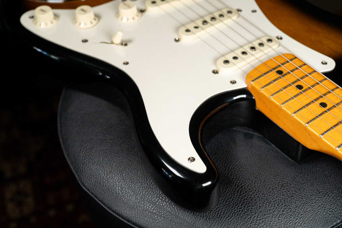 Close-up of a black guitar body with a white pickguard, cream knobs, and a maple fretboard, resting on a black textured surface.