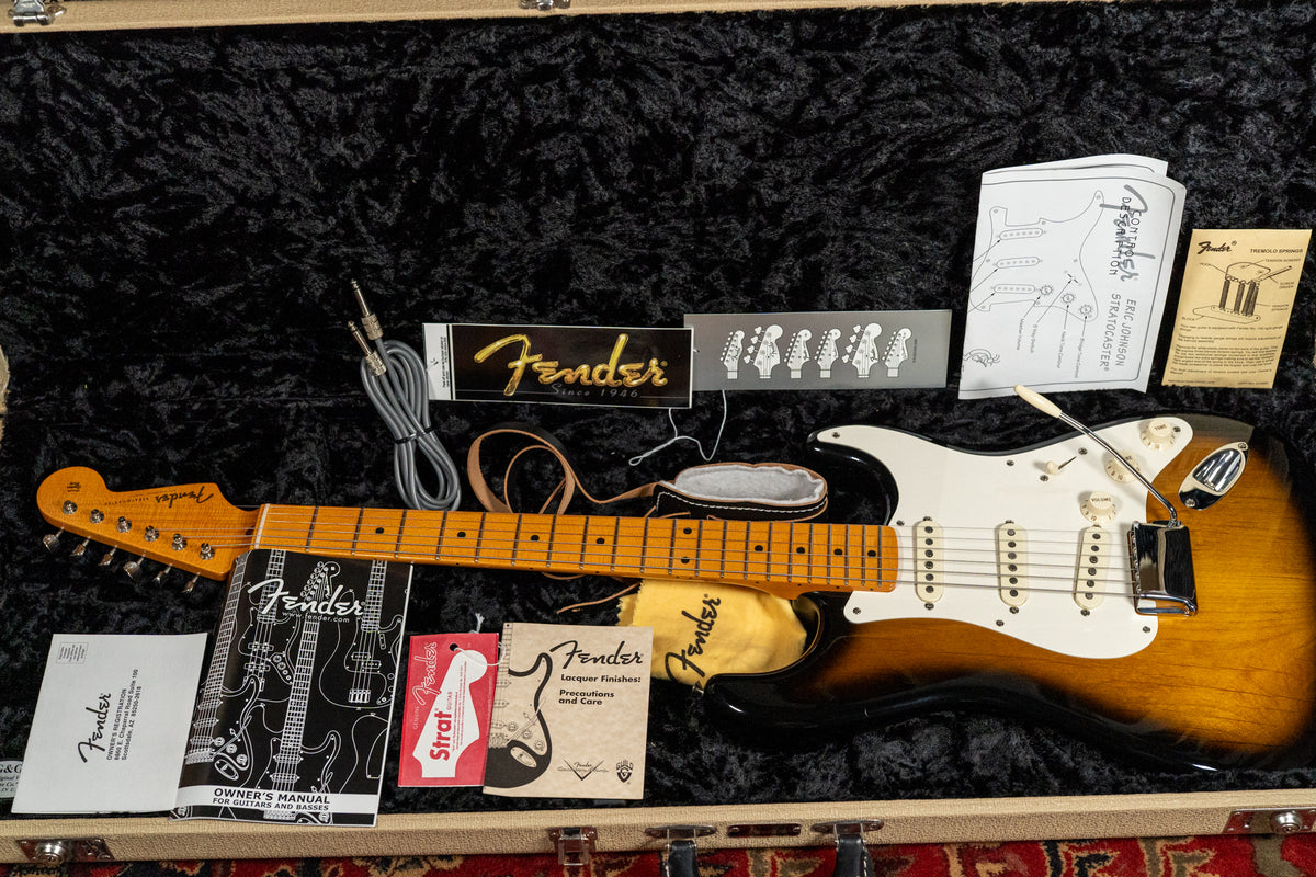 Overhead view of a sunburst Stratocaster in its open case, surrounded by manuals, a cable, and other accessories.