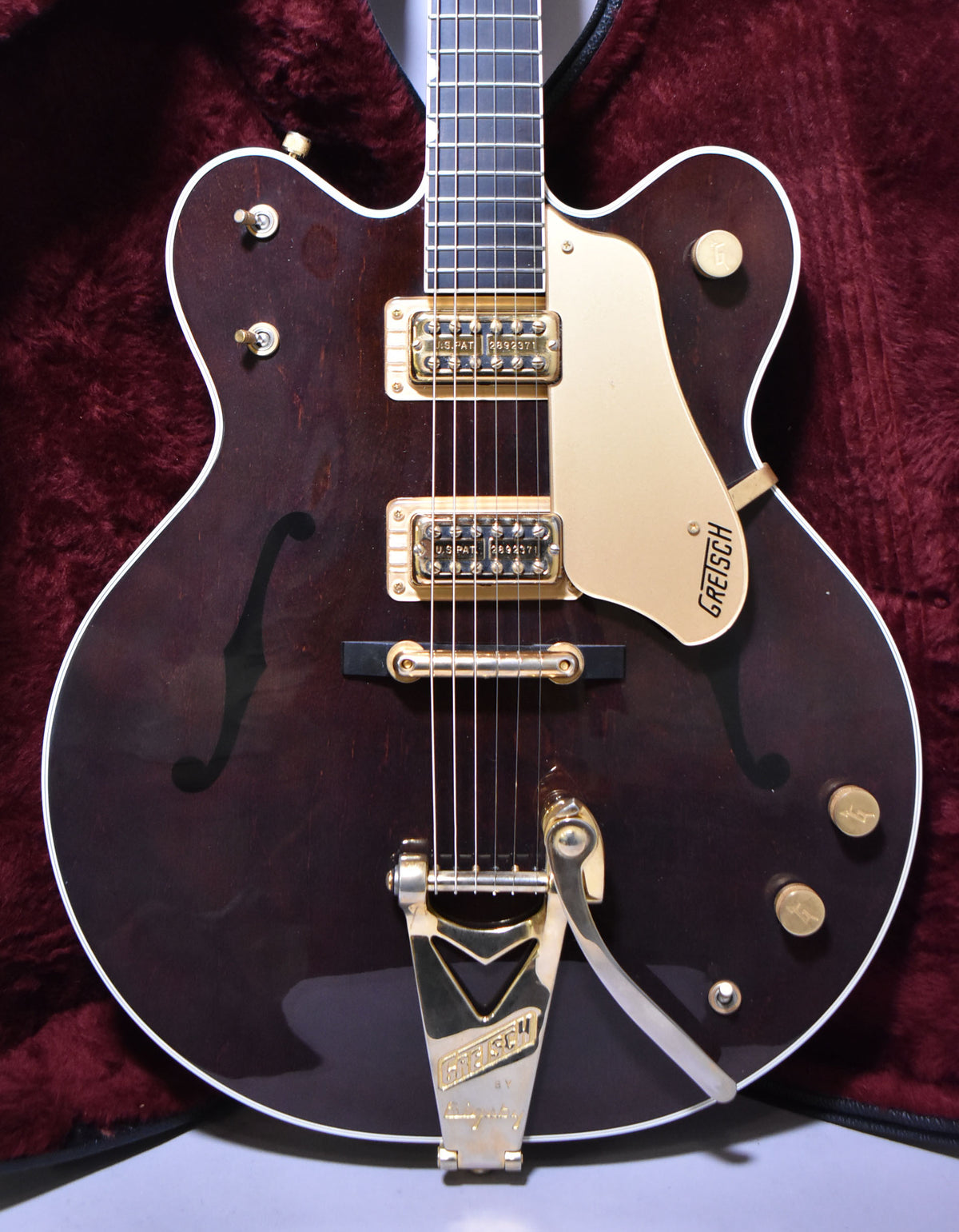 Full-body shot of a dark walnut hollow-body guitar with gold hardware, f-holes, white binding, and a Bigsby tremolo.