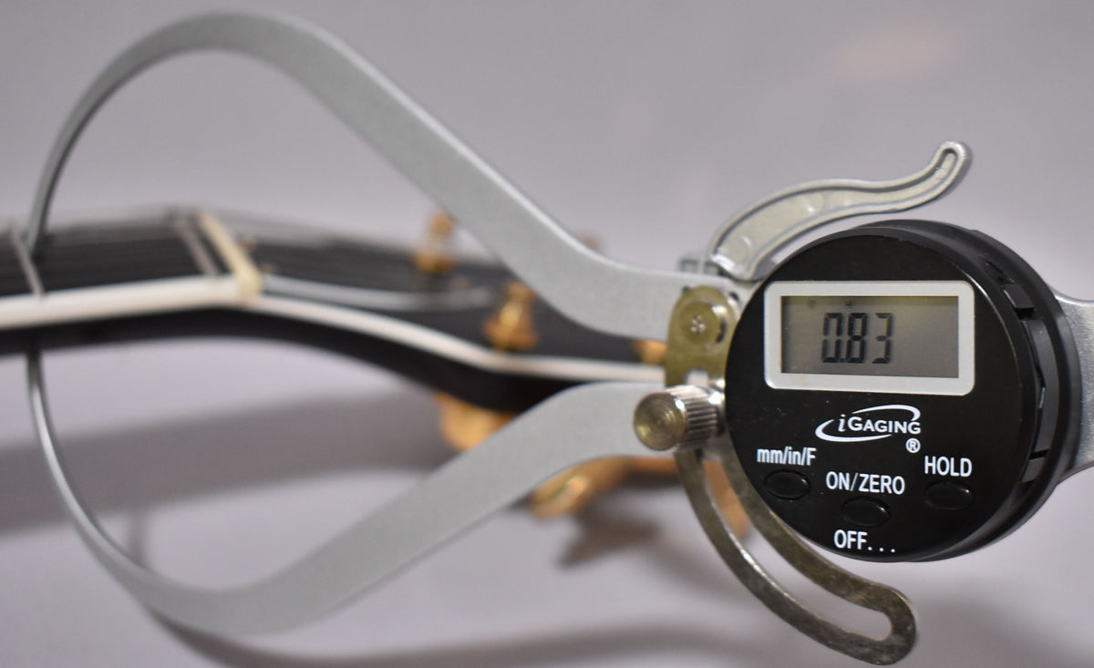 A digital caliper measuring the guitar's nut width, displaying "0.83" on its screen, with the dark headstock visible behind.