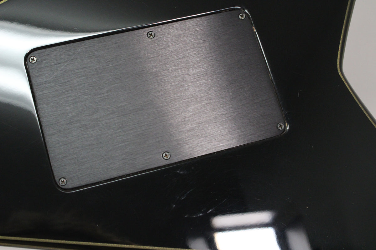 Close-up of the guitar's glossy black body, showing a rectangular brushed metal control cavity cover with four screws and light surface scratches.