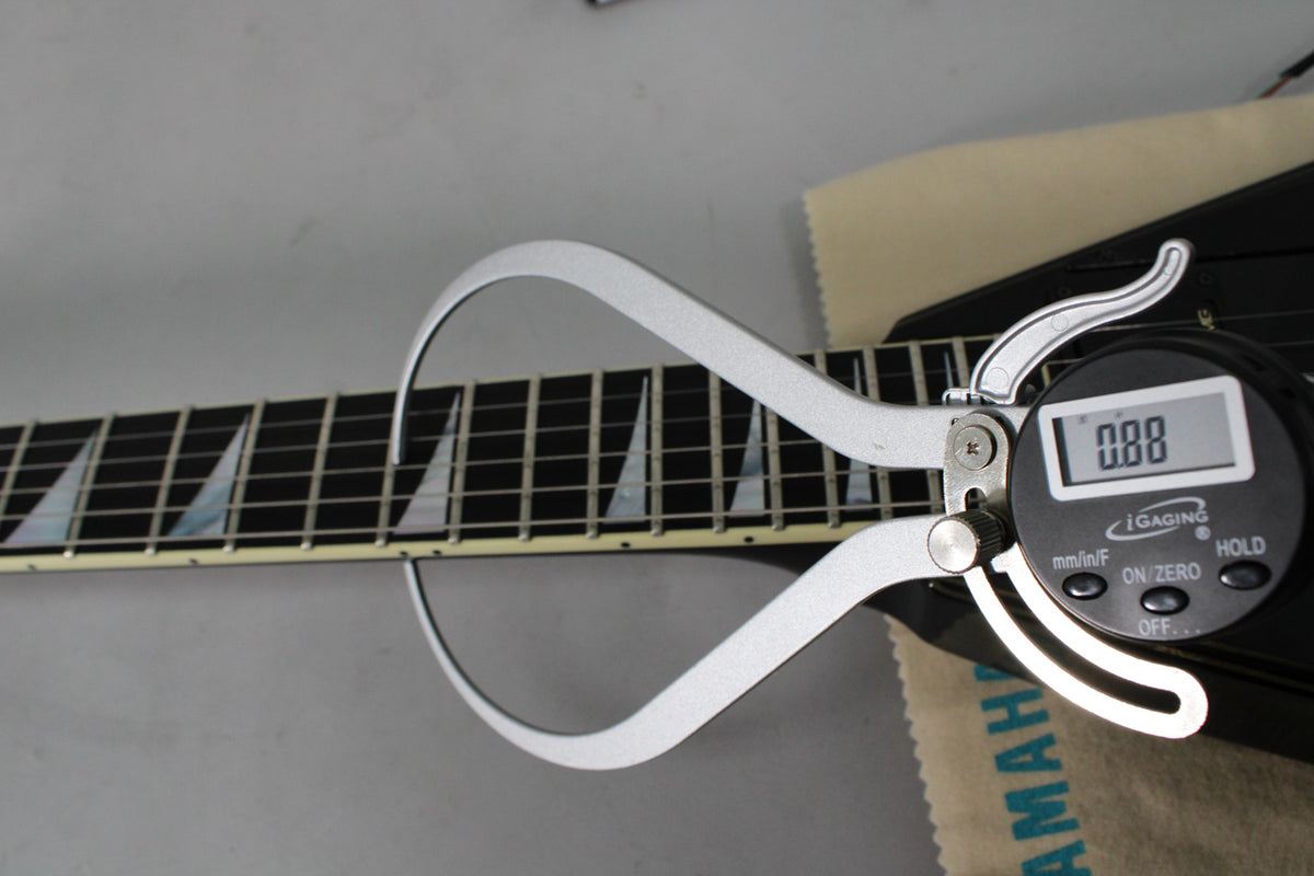 A digital fret caliper measures the black fretboard with pearloid sharkfin inlays on the guitar's neck.