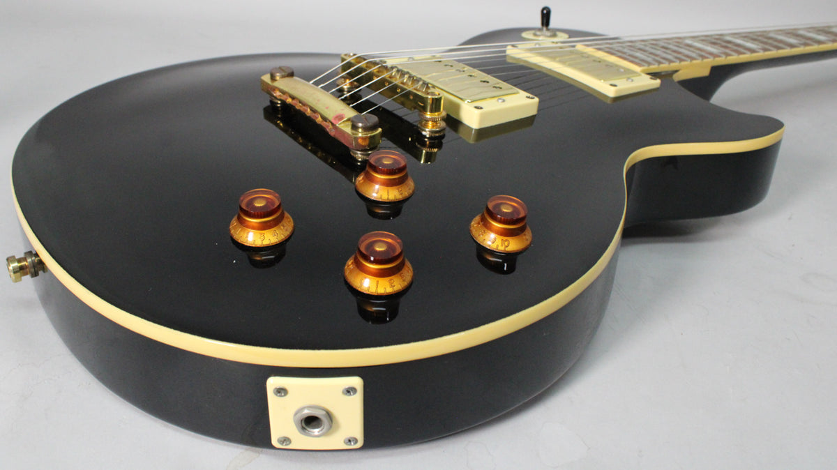 The guitar's glossy black body, with cream binding, shows four amber control knobs and a square cream output jack plate.