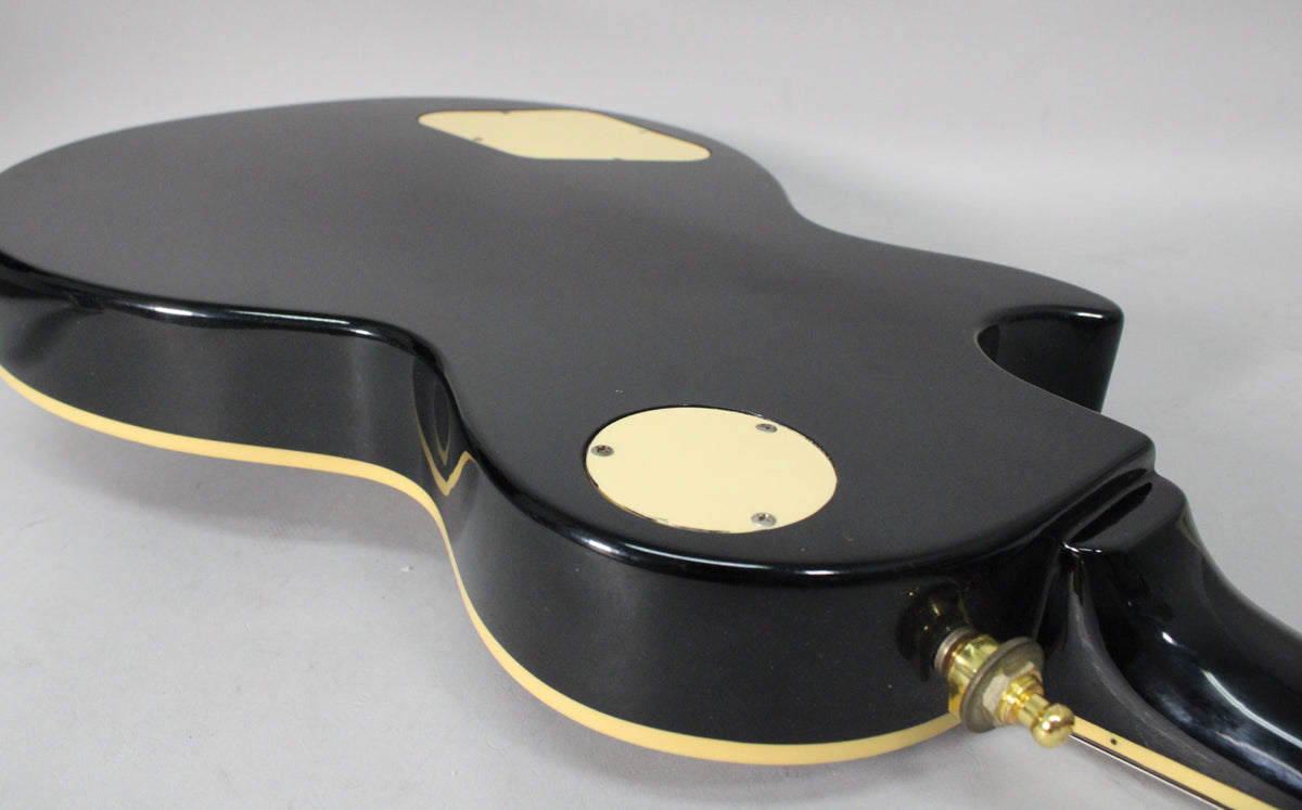 The back of a glossy black guitar body, angled from the lower bout, showing cream binding, control covers, and a gold output jack.