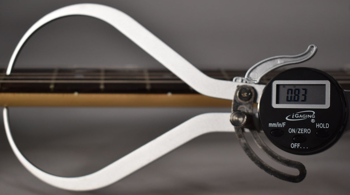 A digital caliper measures the guitar's neck, showing "0.83" on its screen, with the fretboard and strings visible.