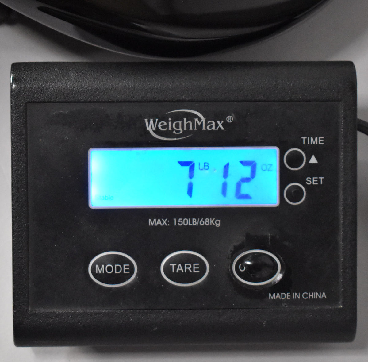A close-up of a black digital scale displaying "7 LB 12 OZ" in bright blue, with "WeighMax" and "MADE IN CHINA" visible.