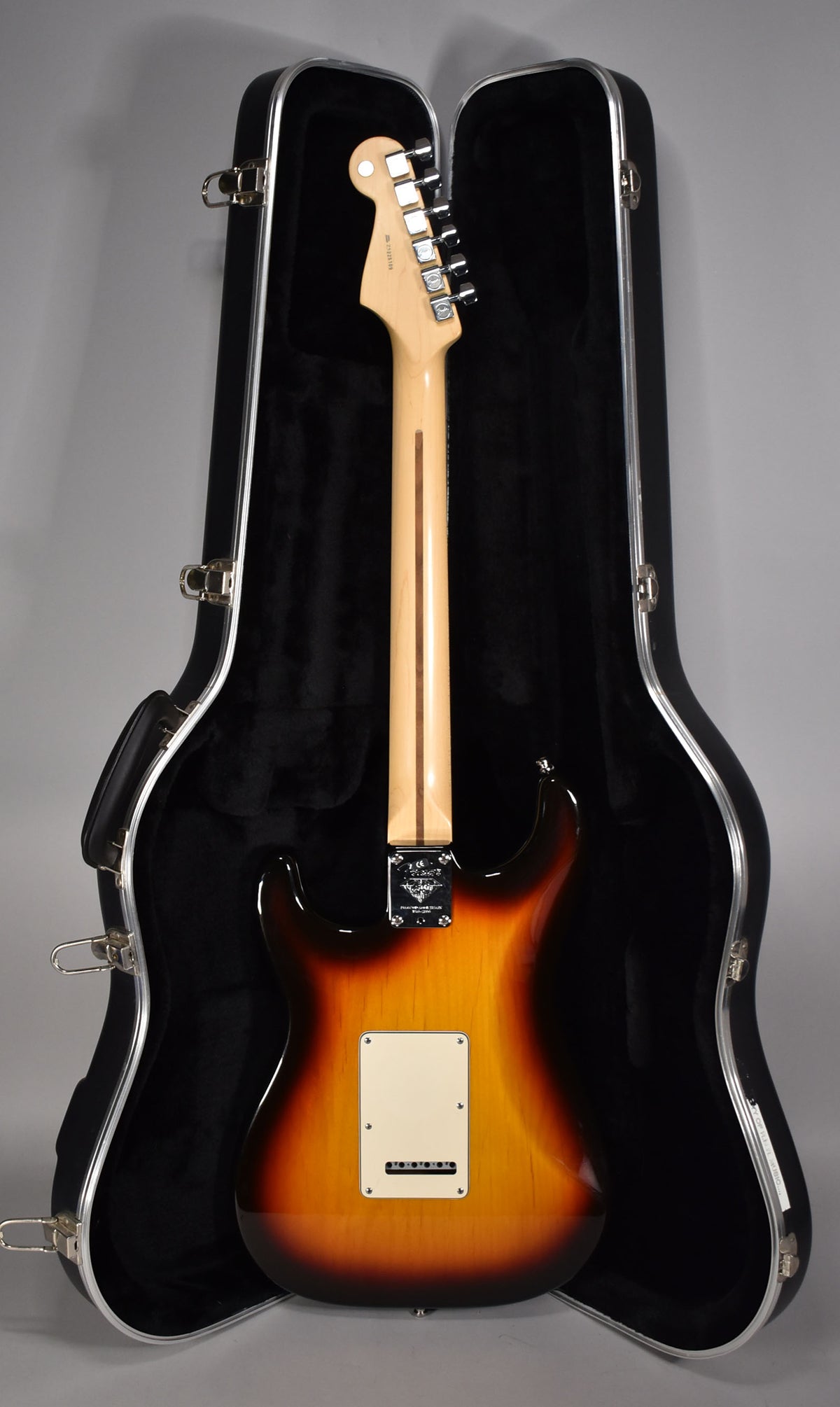 The sunburst Stratocaster, seen from the back, rests in its open hard case, showing its maple neck, chrome tuners, and cream backplate.
