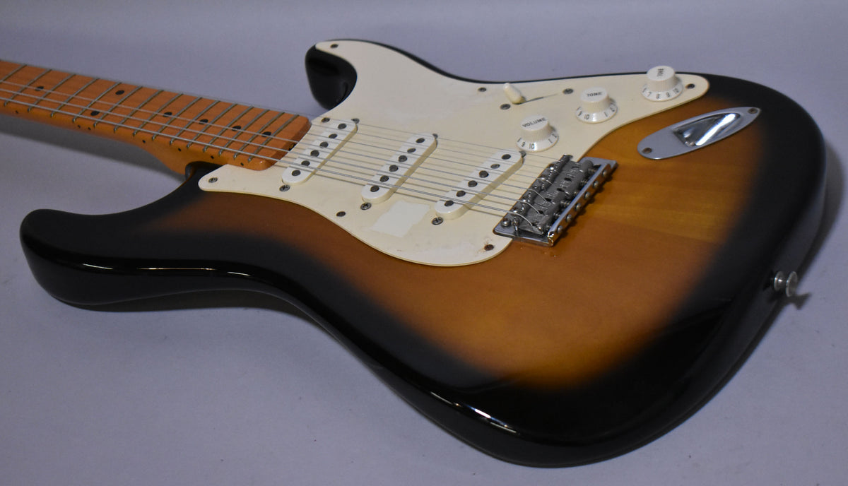 The sunburst guitar body is angled right, showing the maple neck, white pickguard, three single-coil pickups, and chrome hardware.