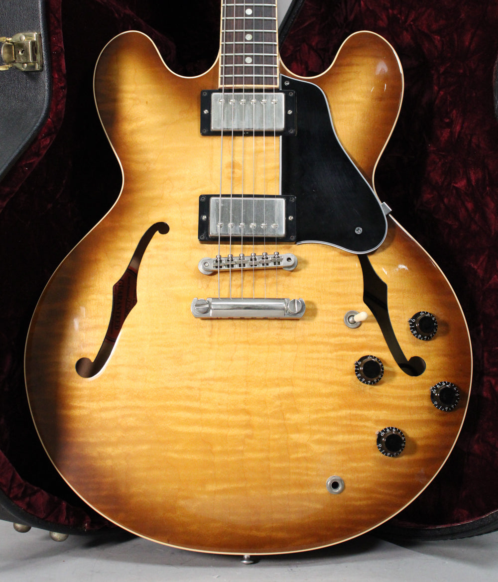 Full front view of a figured sunburst semi-hollow guitar in its red-lined case, showing two humbuckers, black pickguard, and four black knobs.