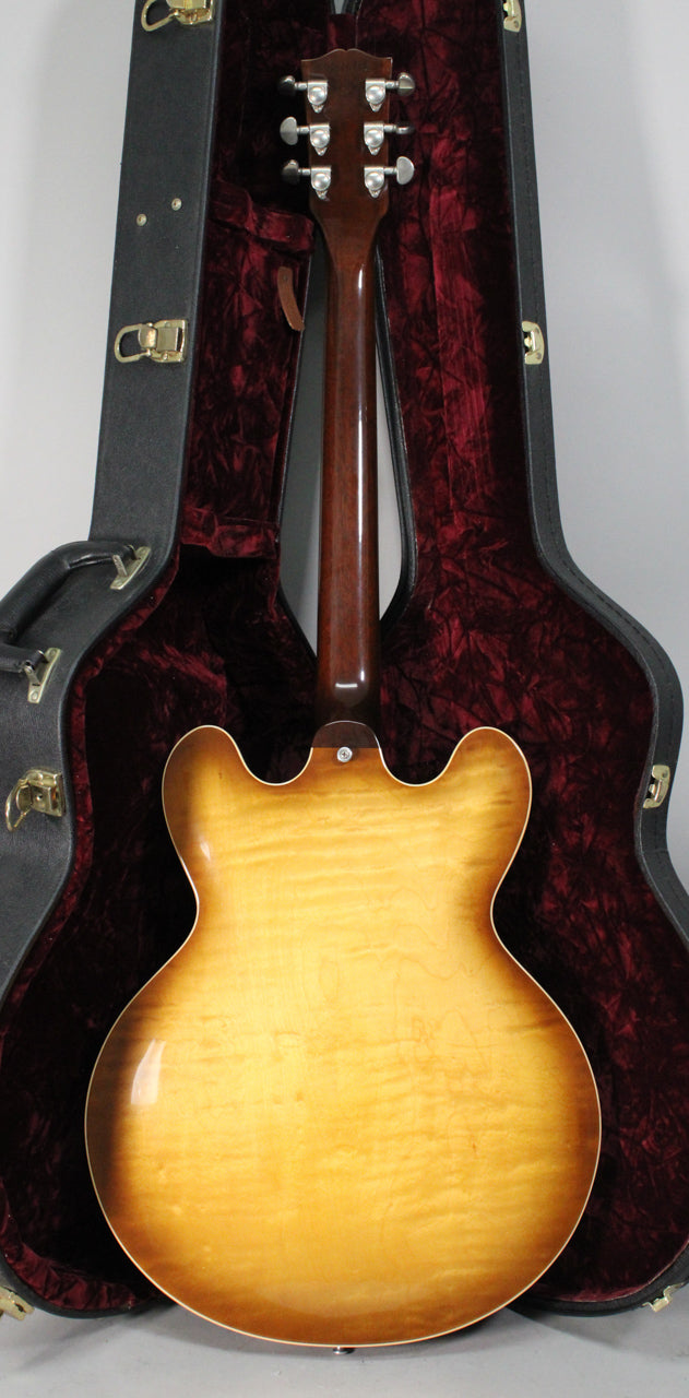 Rear view of a sunburst semi-hollow guitar with figured maple back, dark brown neck, and six chrome tuners, standing in a red-lined case.