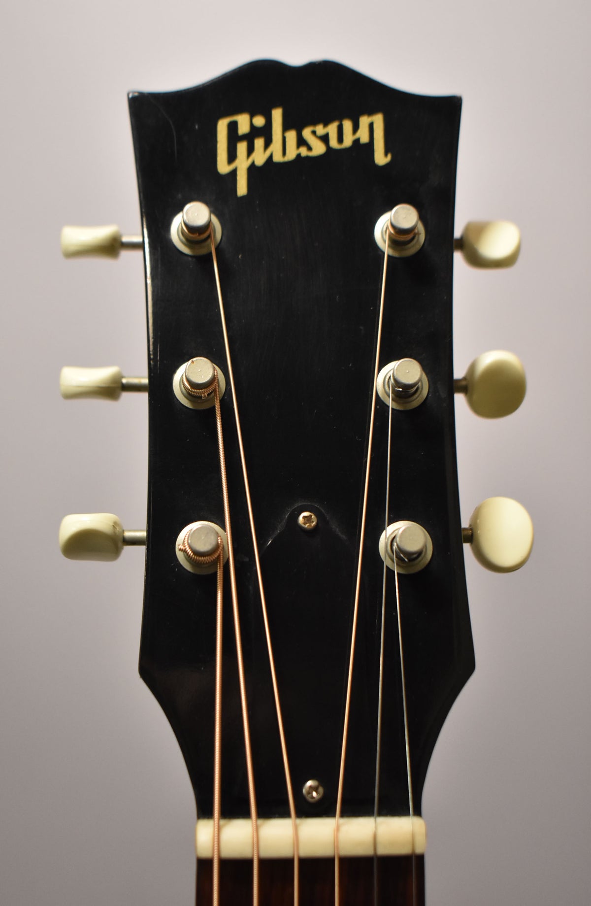 Close-up of the guitar's black headstock with gold lettering, six cream tuning pegs, and light-colored strings.