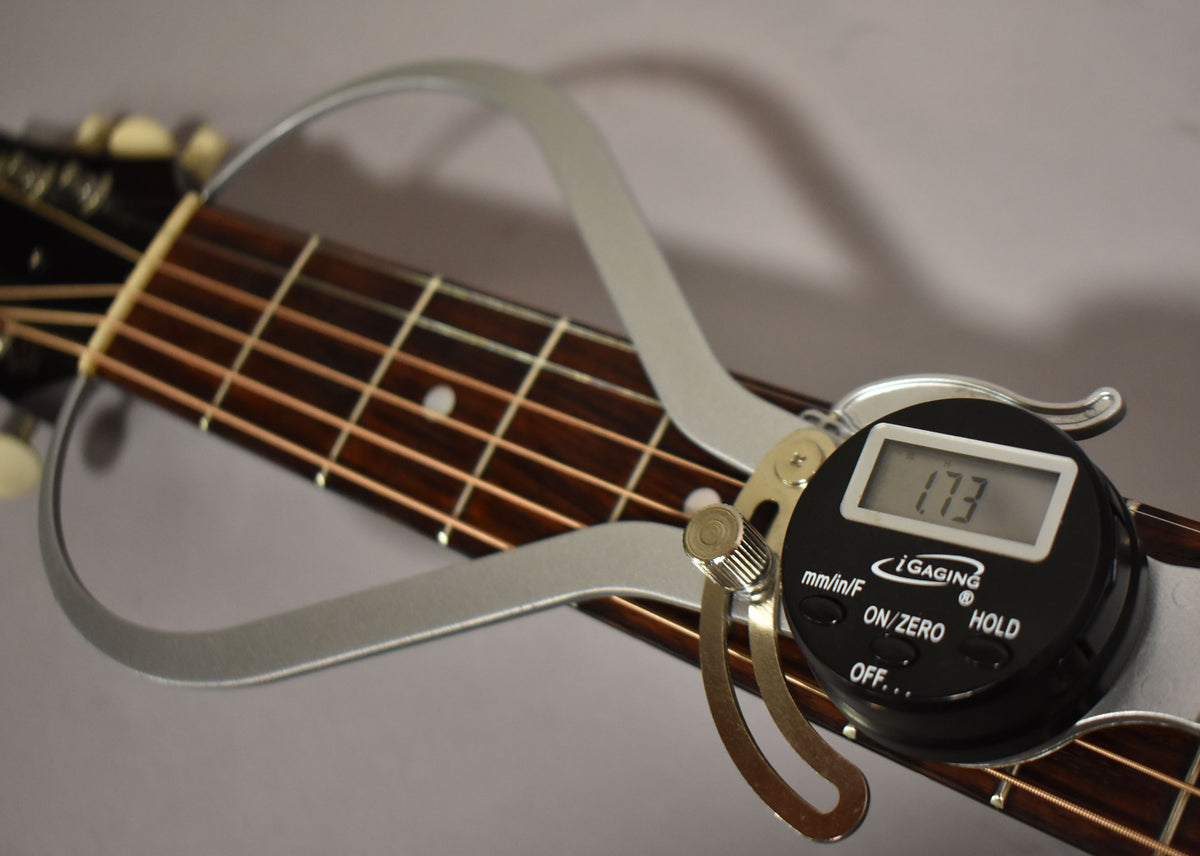 A digital caliper measures the string height at the first fret of the guitar's dark rosewood fretboard.