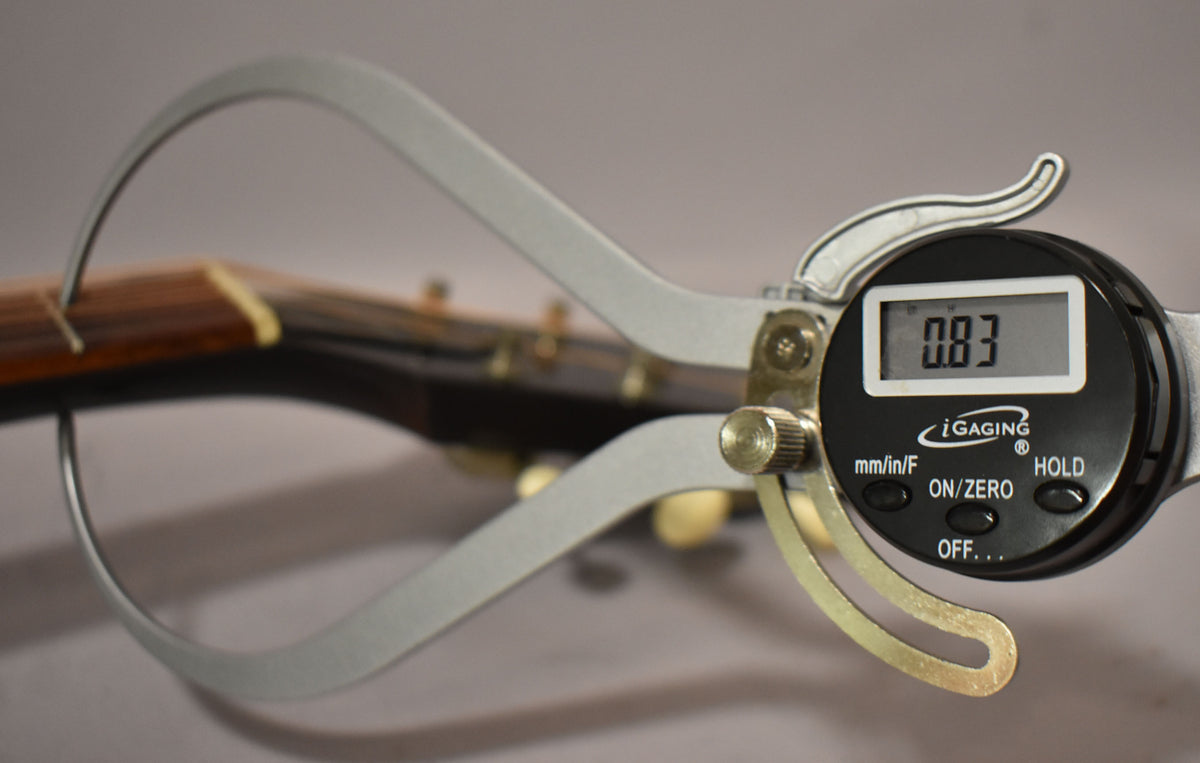 A digital caliper measures the string height at the first fret, displaying 0.83mm, with the headstock blurred in the background.