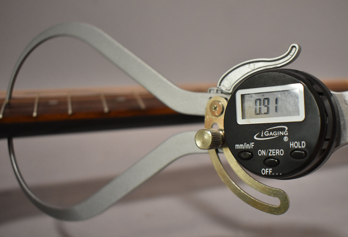 A digital caliper measures the string height at the 12th fret, displaying "0.91" on its screen.