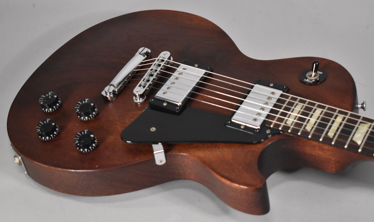 Angled close-up of a dark brown electric guitar body with a satin finish, showing chrome hardware, black pickguard, and four black knobs.