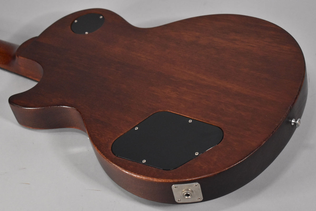 The back of a faded walnut guitar shows a black control cavity cover and output jack, with light wear.