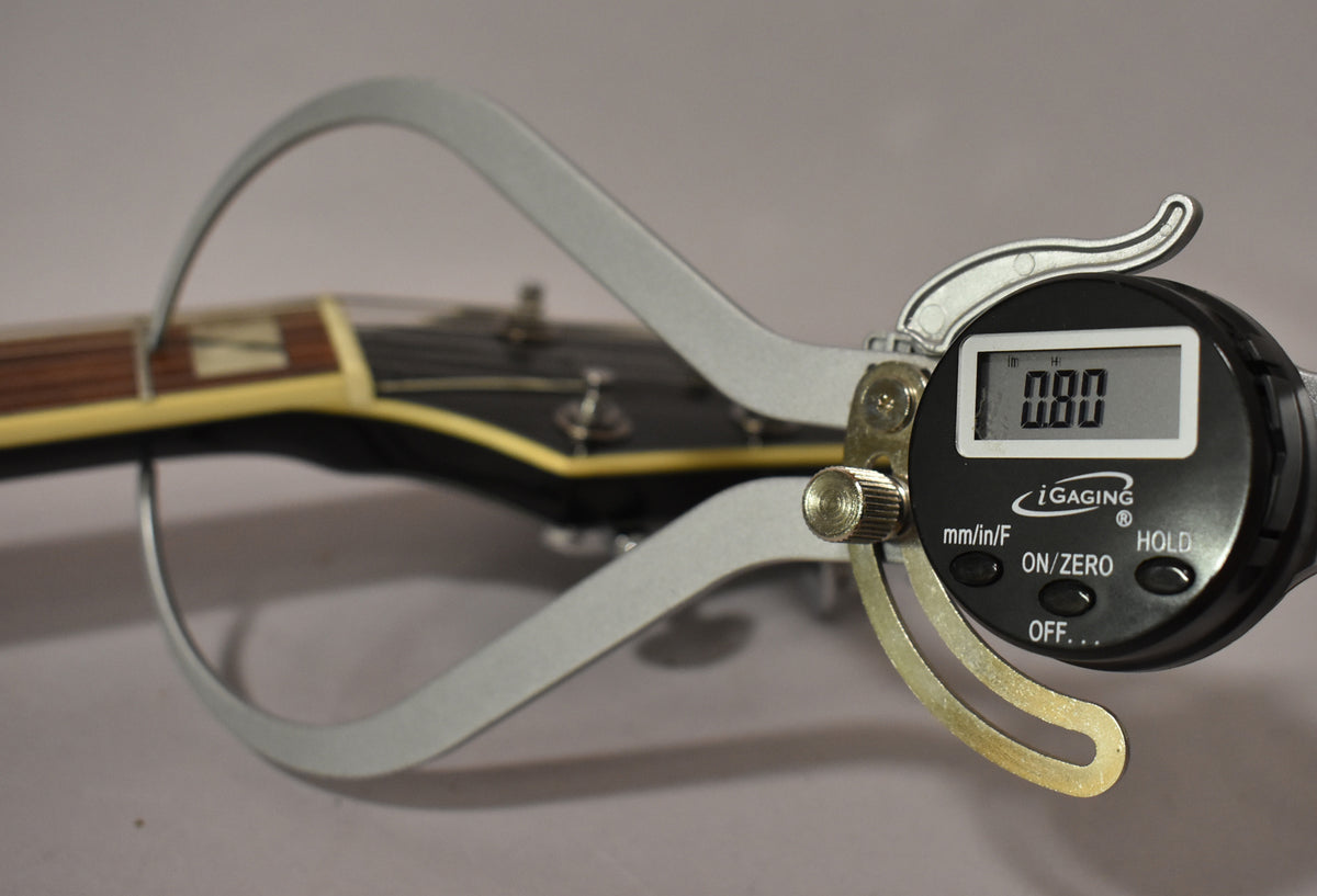 A digital caliper measures the neck thickness of the guitar, showing 0.80 on its display, with the headstock blurred in the background.